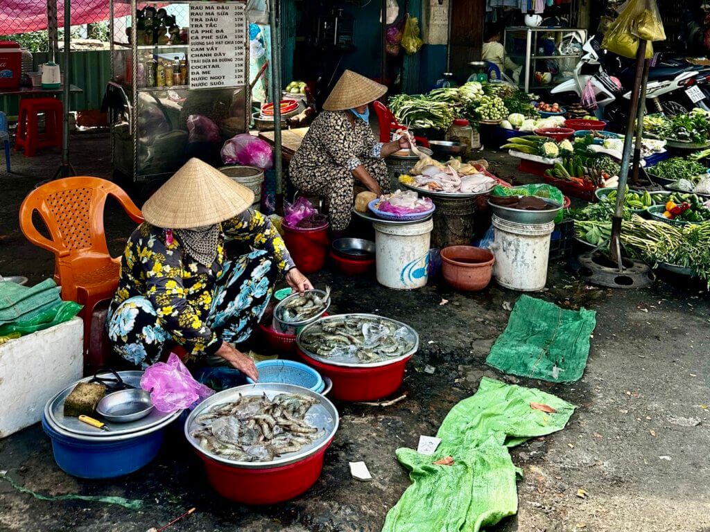Straßenmarkt in Ho-Chi-Minh-Stadt mit Verkäuferinnen in traditionellen Hüten, frischem Gemüse und Meeresfrüchten