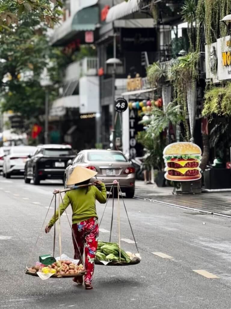 Straßenszene in Hue, Vietnam: Frau mit Gemüse im Schulterkorb läuft an Fast-Food-Werbung vorbei – Alltag während einer 4 Wochen Vietnam Reise