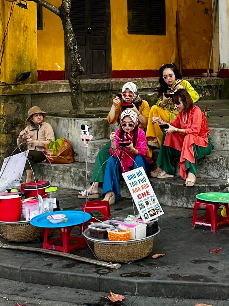 Straßenszene in Hoi An, Vietnam: Frauen in bunten traditionellen Kleidern essen Streetfood an einem Marktstand in der Altstadt