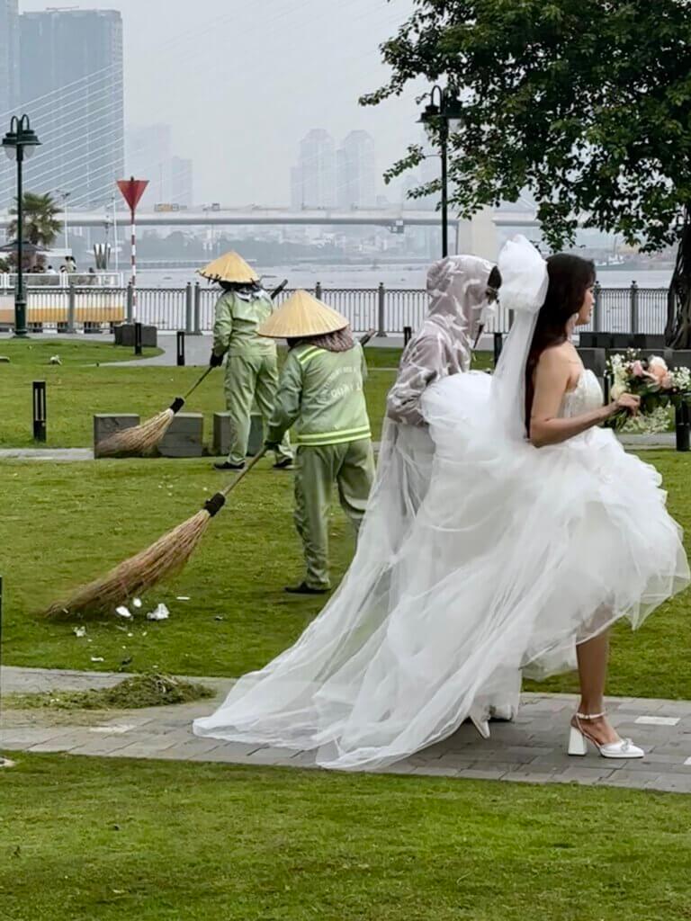 Szene am Flussufer in Saigon, Vietnam: Braut im weißen Kleid neben Straßenkehrern – Momentaufnahme während einer 4 Wochen Vietnam Reise