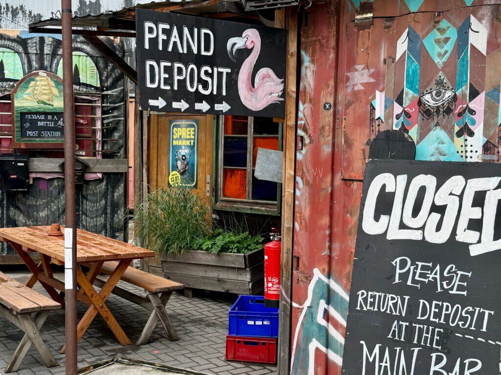 Szene aus dem alternativen Kulturareal Holzmarkt Berlin mit Pfandstation, bemalten Containern, Wandkunst und Hinweisschildern in bunter Umgebung