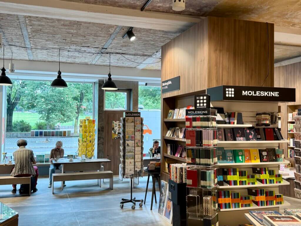Innenansicht der Ocelot-Buchhandlung mit Leseplätzen vor großen Fenstern, Moleskine-Display und Menschen beim Schmökern in Berlin-Mitte
