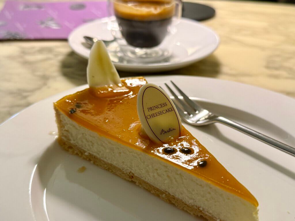 Cheesecake mit Passionsfrucht und weißer Schokolade auf weißem Teller im Café Princess Cheesecake, Berlin-Mitte, mit Espresso im Hintergrund