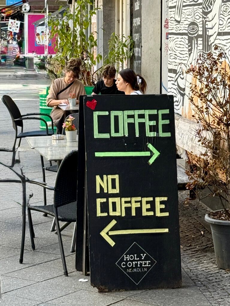 Schild vor dem Café „Holy Coffee“ in Berlin-Neukölln mit der Aufschrift „COFFEE → / NO COFFEE ←“, drei Gäste sitzen im Außenbereich.