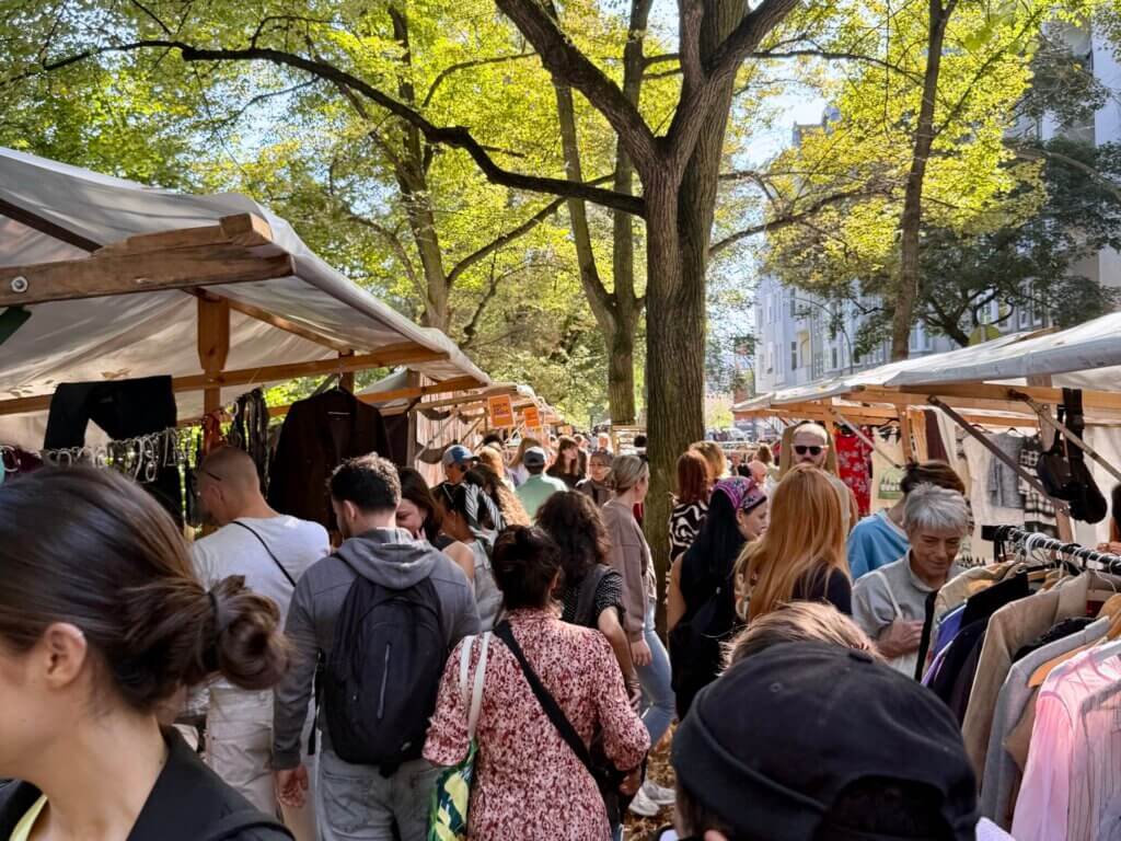 Menschenmengen auf dem Nowkoelln Flowmarkt in Berlin-Neukölln, zwischen Marktständen unter alten Bäumen.