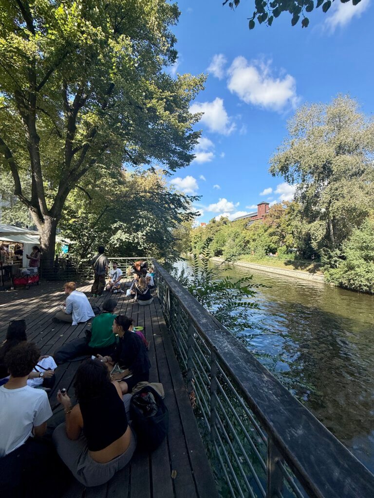 Menschen sitzen entspannt auf einem Holzdeck am Maybachufer in Berlin, mit Blick auf den Kanal.
