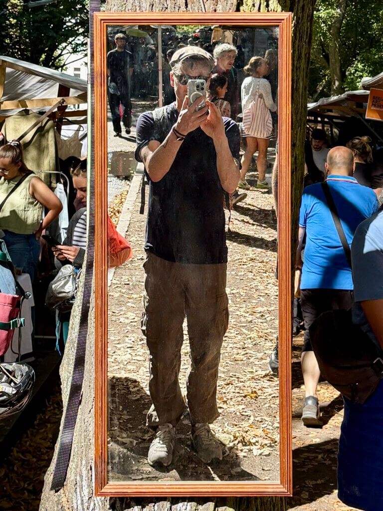 Spiegelbild eines Mannes auf dem Nowkoelln Flowmarkt in Berlin-Neukölln, umgeben von Flohmarktständen und Menschen unter Bäumen