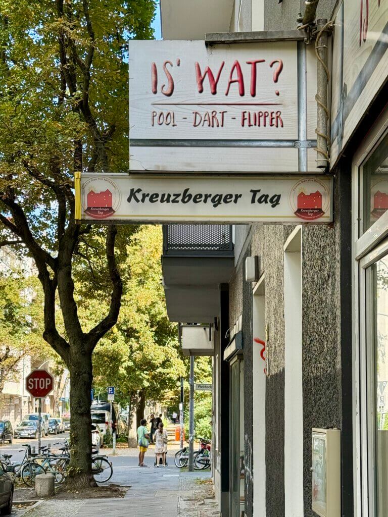 Schild mit der Aufschrift „Is’ Wat?“ über einer Berliner Bar in der Nähe der Weserstraße, darunter weiteres Schild „Kreuzberger Tag“, Kiezszene mit Bäumen und Fußgängern.