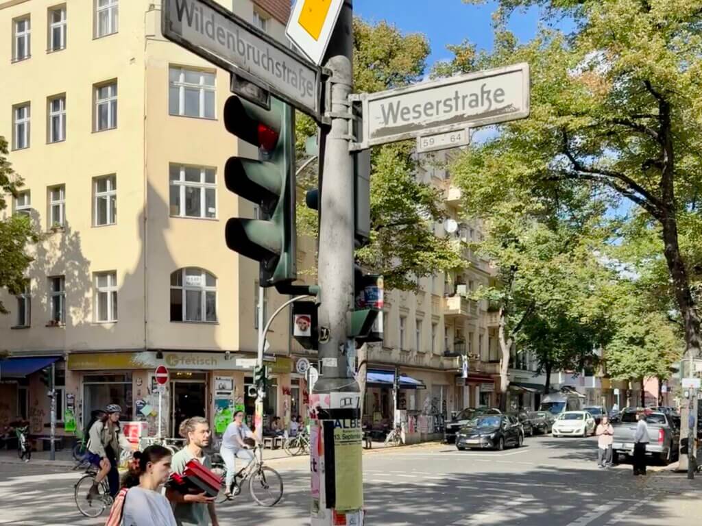 Straßenschilder an der Kreuzung Weserstraße und Wildenbruchstraße in Berlin-Neukölln, Menschen zu Fuß und mit dem Fahrrad unterwegs, Kiezszene bei Sonne.