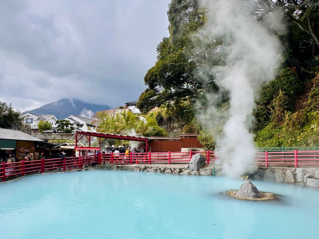 Heiße Quelle in Beppu mit hellblauem Wasser, aufsteigendem Dampf und roten Geländern, im Hintergrund Berge und Besuchergruppen.