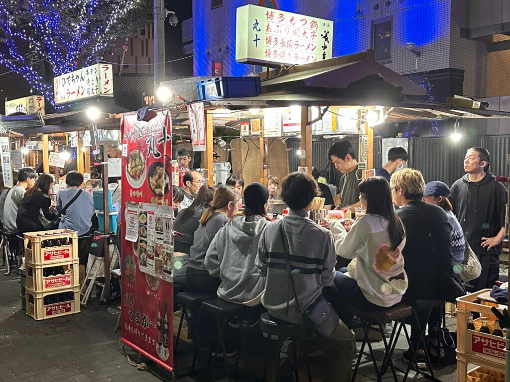 Menschen sitzen an beleuchteten Food Stalls in Fukuoka bei Nacht, umgeben von Schildern und buntem Treiben.