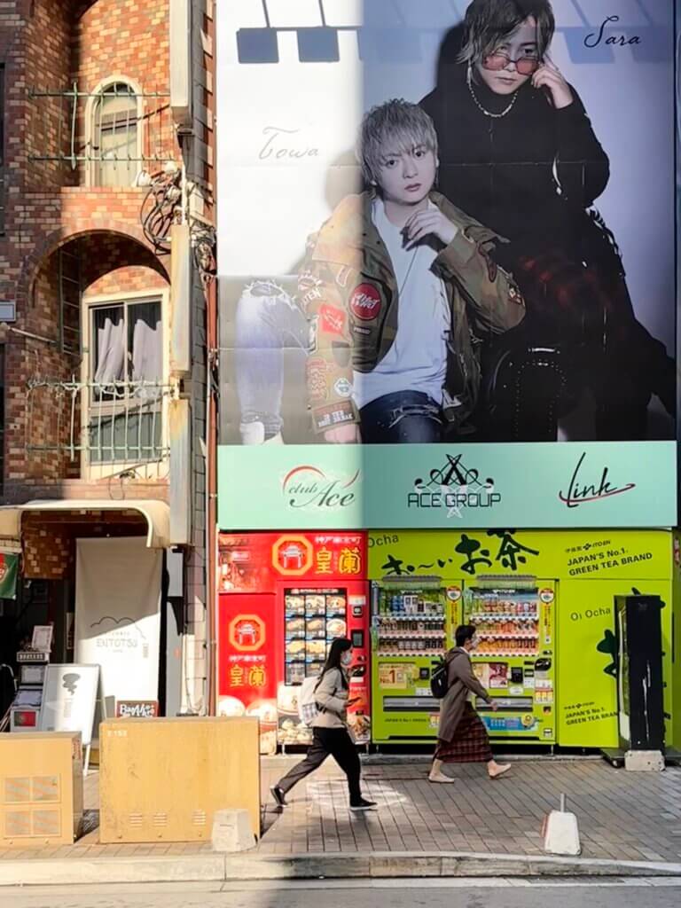 Straßenszene im Stadtteil Sannomiya in Kobe mit großen Werbeplakaten, bunten Getränkeautomaten und vorbeilaufenden Passanten im Vormittagslicht.