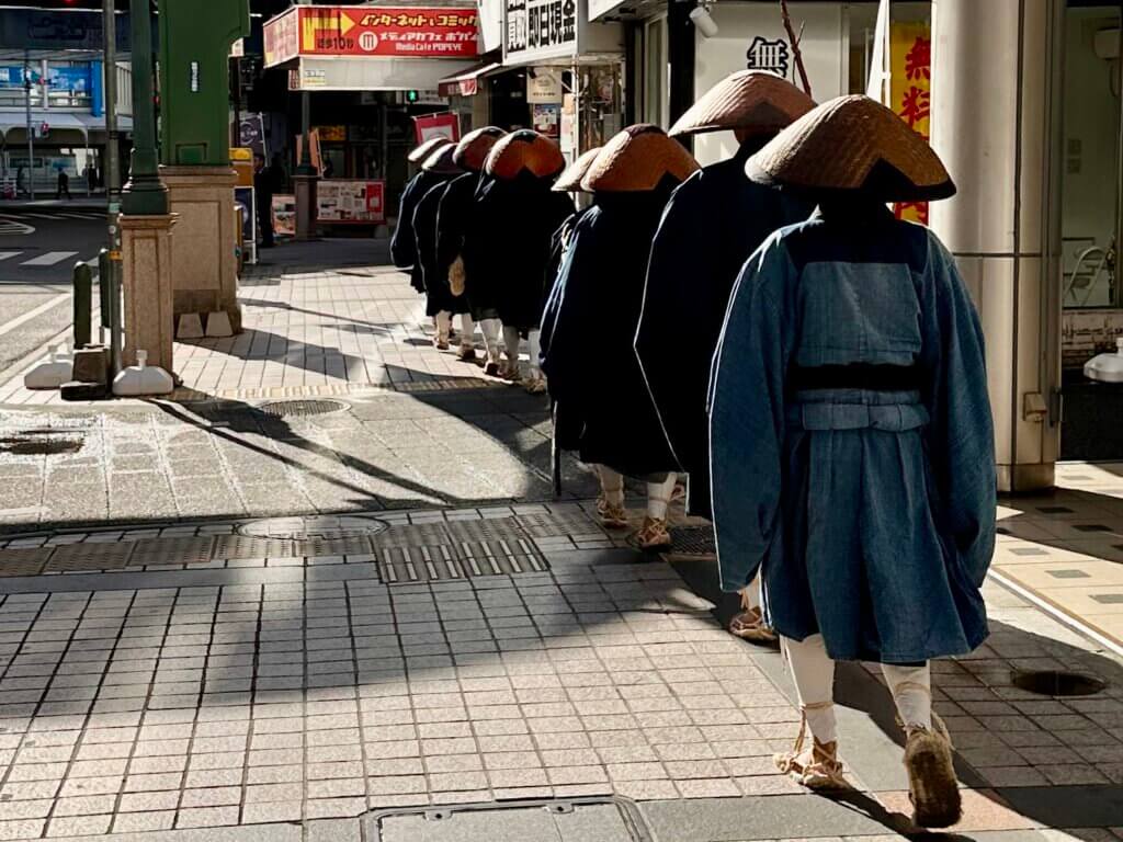 Eine Gruppe traditionell gekleideter Mönche mit Strohhüten geht in Reihe durch eine moderne Einkaufsstraße in Kobe, flankiert von Geschäften und Reklame.