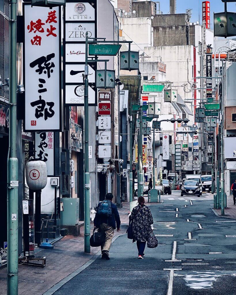 Zwei Menschen mit Taschen laufen durch eine Straße im Zentrum von Kumamoto, flankiert von japanischen Schildern und Bars.