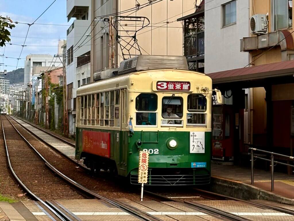 Alte Straßenbahn in grün-gelber Lackierung fährt durch ein Wohnviertel in Nagasaki bei Tageslicht.