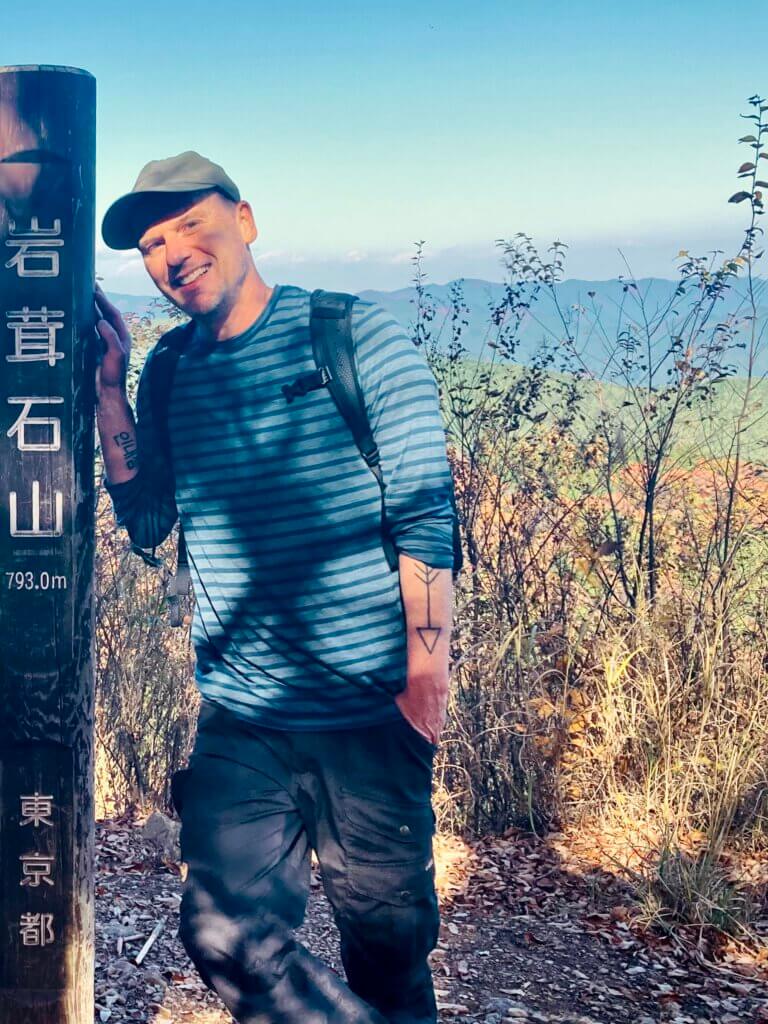 Person mit Rucksack lehnt an einem hölzernen Wegweiser auf einem Berg in Ōme bei Tokio, umgeben von herbstlicher Landschaft.