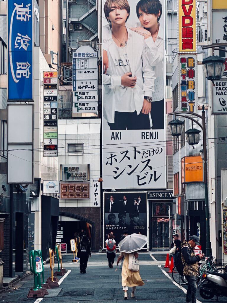 Straßenszene im Tokioter Stadtteil Shinjuku mit großen Werbetafeln, Regenschirmträgerin und morgendlicher Stimmung.