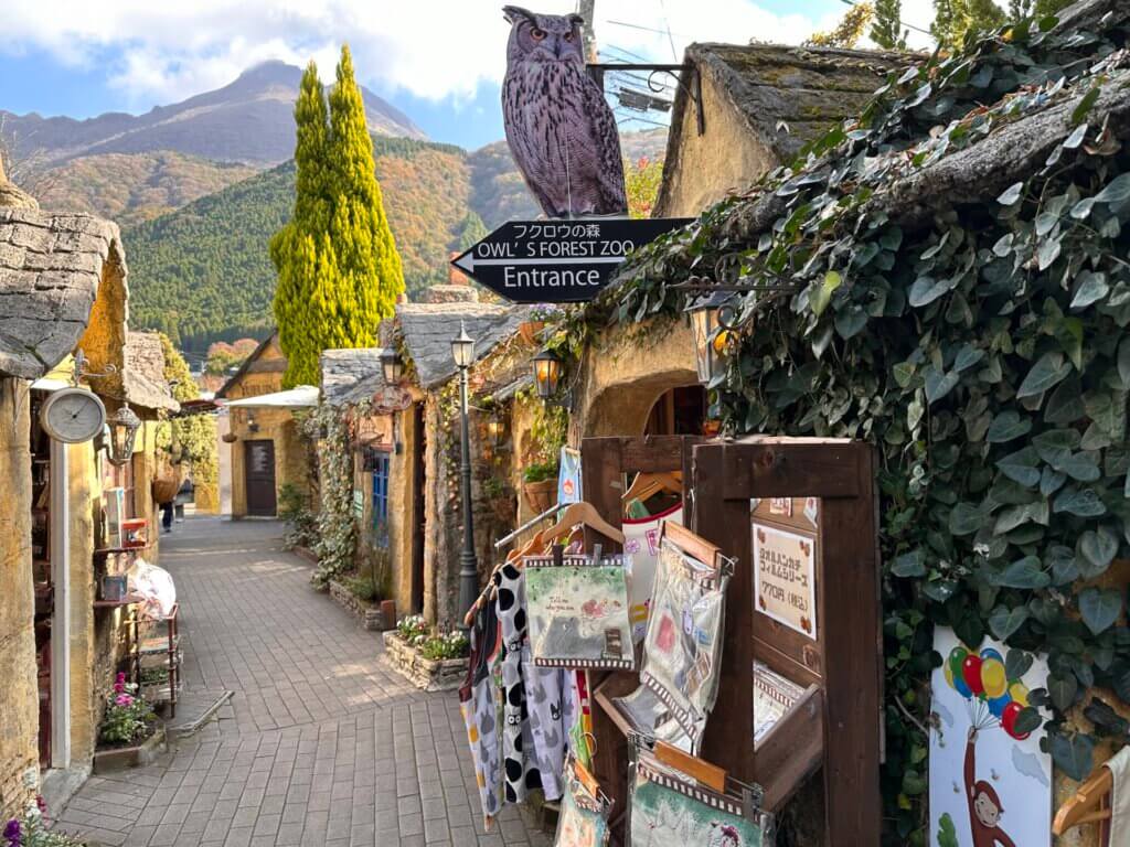Bunte Gassen im Floral Village in Yufuin mit Märchenhäusern, Eulen-Zoo und Blick auf den Berg im Hintergrund.