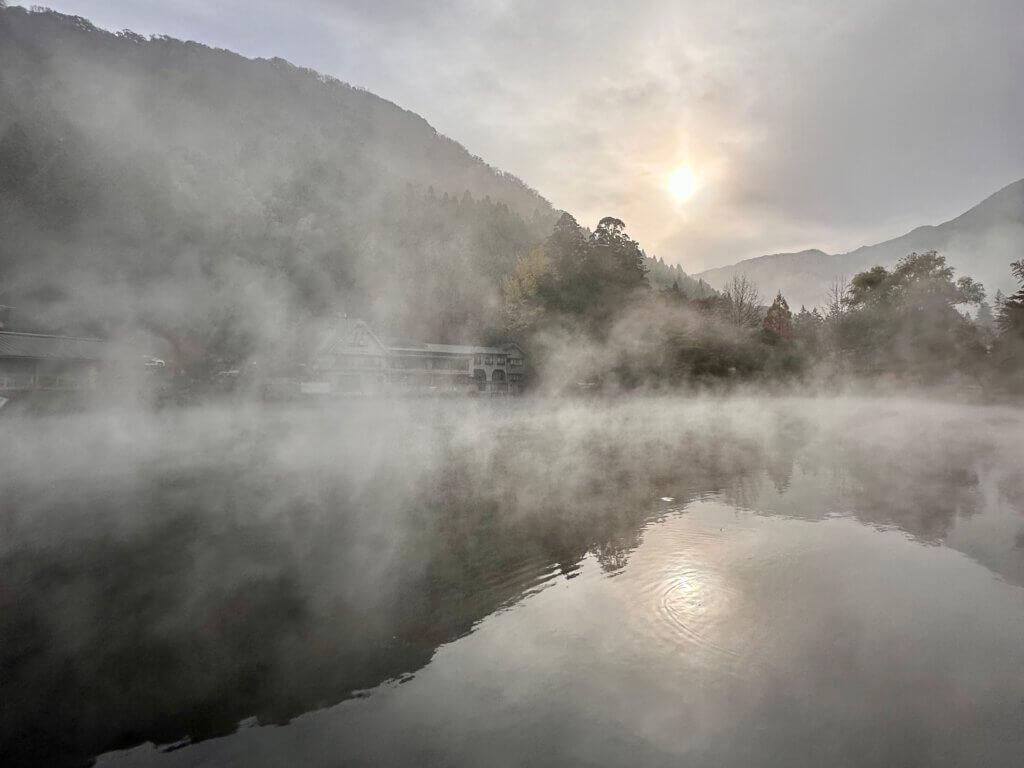 Nebel liegt über dem Lake Kinrin in Yufuin bei Sonnenaufgang, die Berge spiegeln sich im ruhigen Wasser.