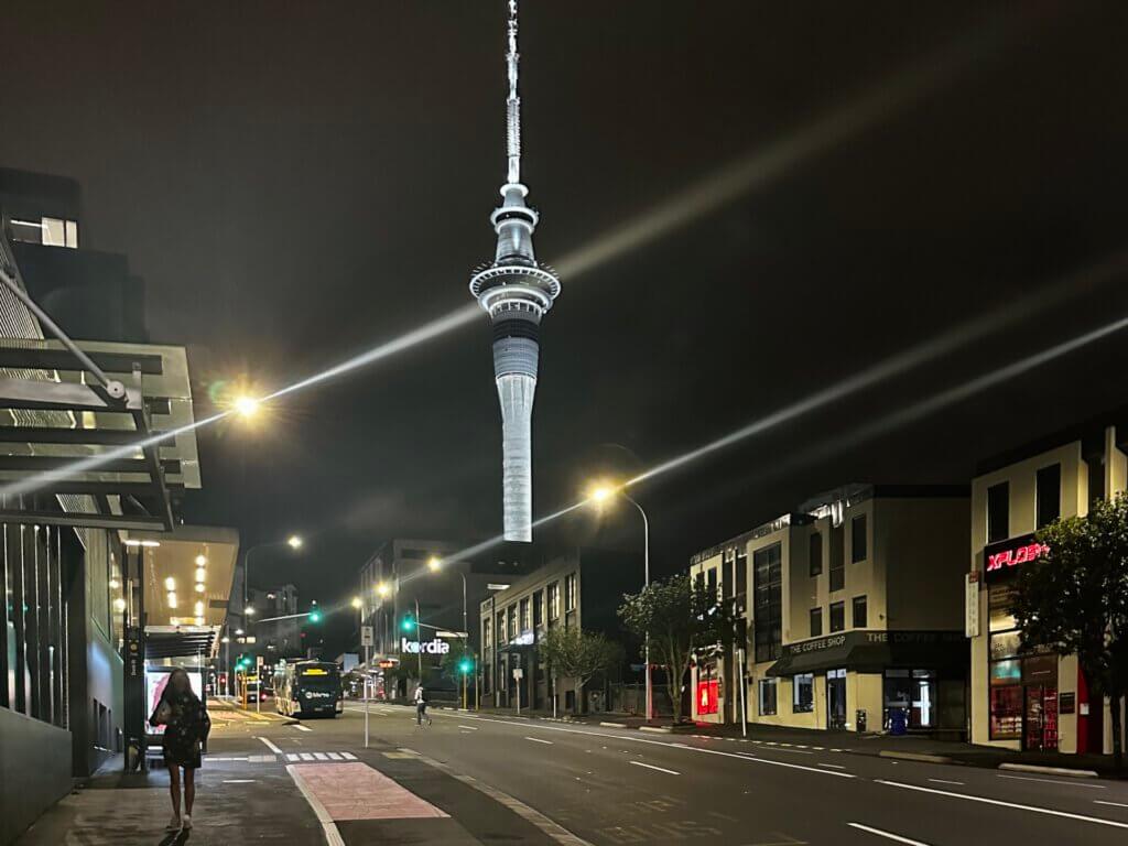 Aucklands Sky Tower bei Nacht, angestrahlt und von leeren Straßen umgeben, mit vereinzelten Fußgängern und beleuchteten Gebäuden.