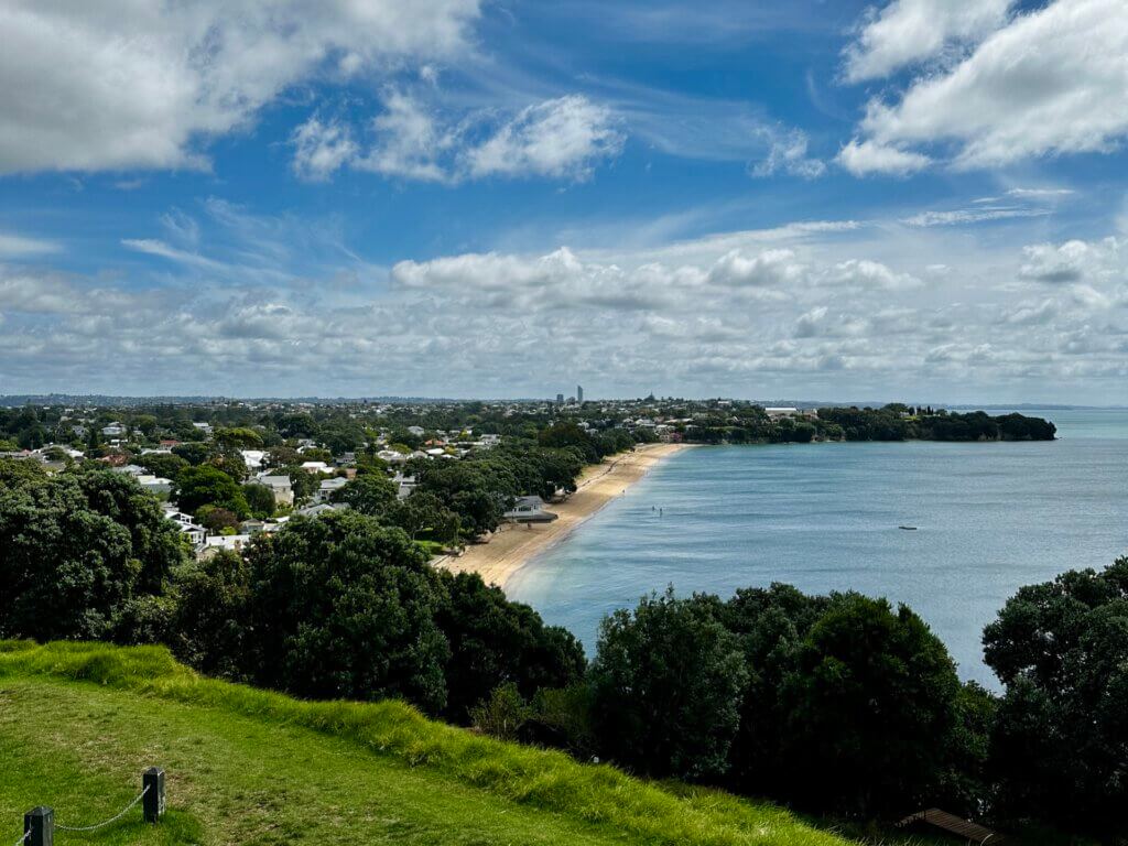 Blick vom North Head in Devonport auf die langgestreckte Küste, den Strand und das dahinterliegende Wohnviertel bei leicht bewölktem Himmel.