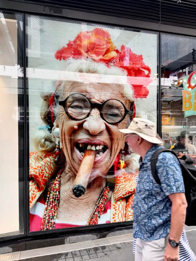 Straßenszene in Auckland: Ein Mann mit Hut läuft an einem farbenfrohen Schaufensterbild einer kubanischen Frau mit Zigarre vorbei
