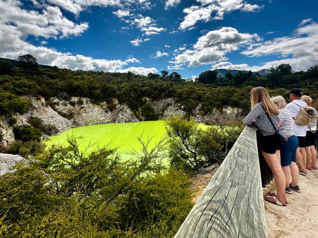 Grün leuchtender Schwefelsee („Devil’s Bath“) im Wai-O-Tapu Thermal Wonderland nahe Rotorua, umgeben von Wald und Felsen; Besucher:innen schauen vom Holzsteg aus auf das surreale Farbenspiel.