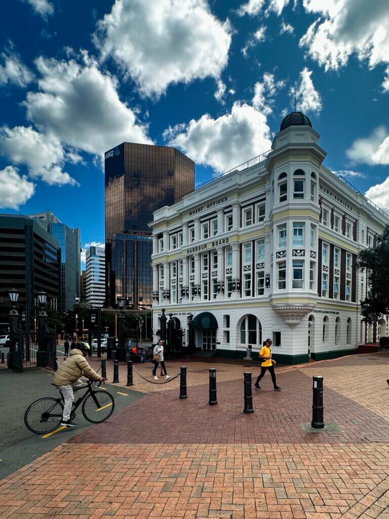 Historisches Gebäude am Wellington Waterfront Walk mit moderner Skyline im Hintergrund, Fußgänger:innen und Radfahrer:innen in der Innenstadt von Neuseelands Hauptstadt.