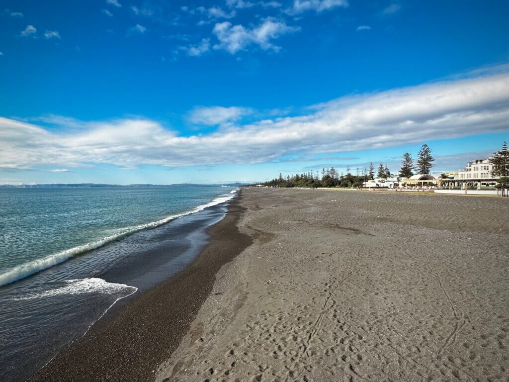 Weitläufiger schwarzer Sandstrand in Napier (Marine Parade Beach) mit ruhiger Brandung und Blick auf das Meer unter blauem Himmel.
